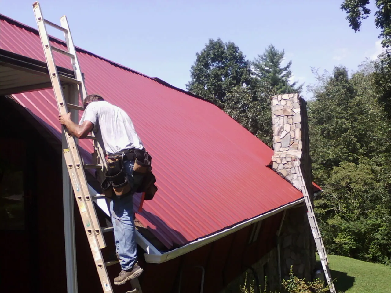 Roofer on a ladder beside a red metal roof and stone chimney — Soffit & Fascia Repair by Roofing Brothers in Trumansburg